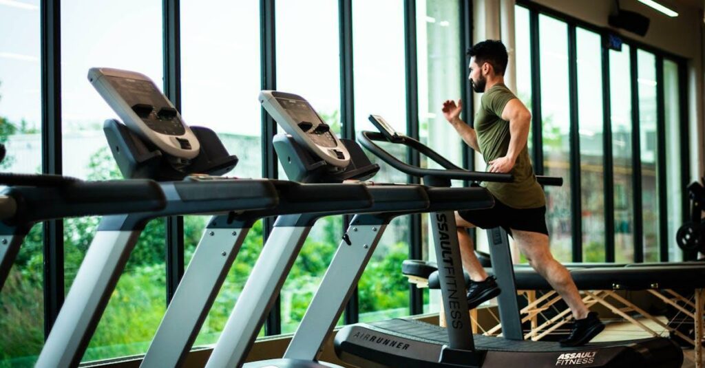 Young man workouts on treadmill in modern gym with large windows and natural light.